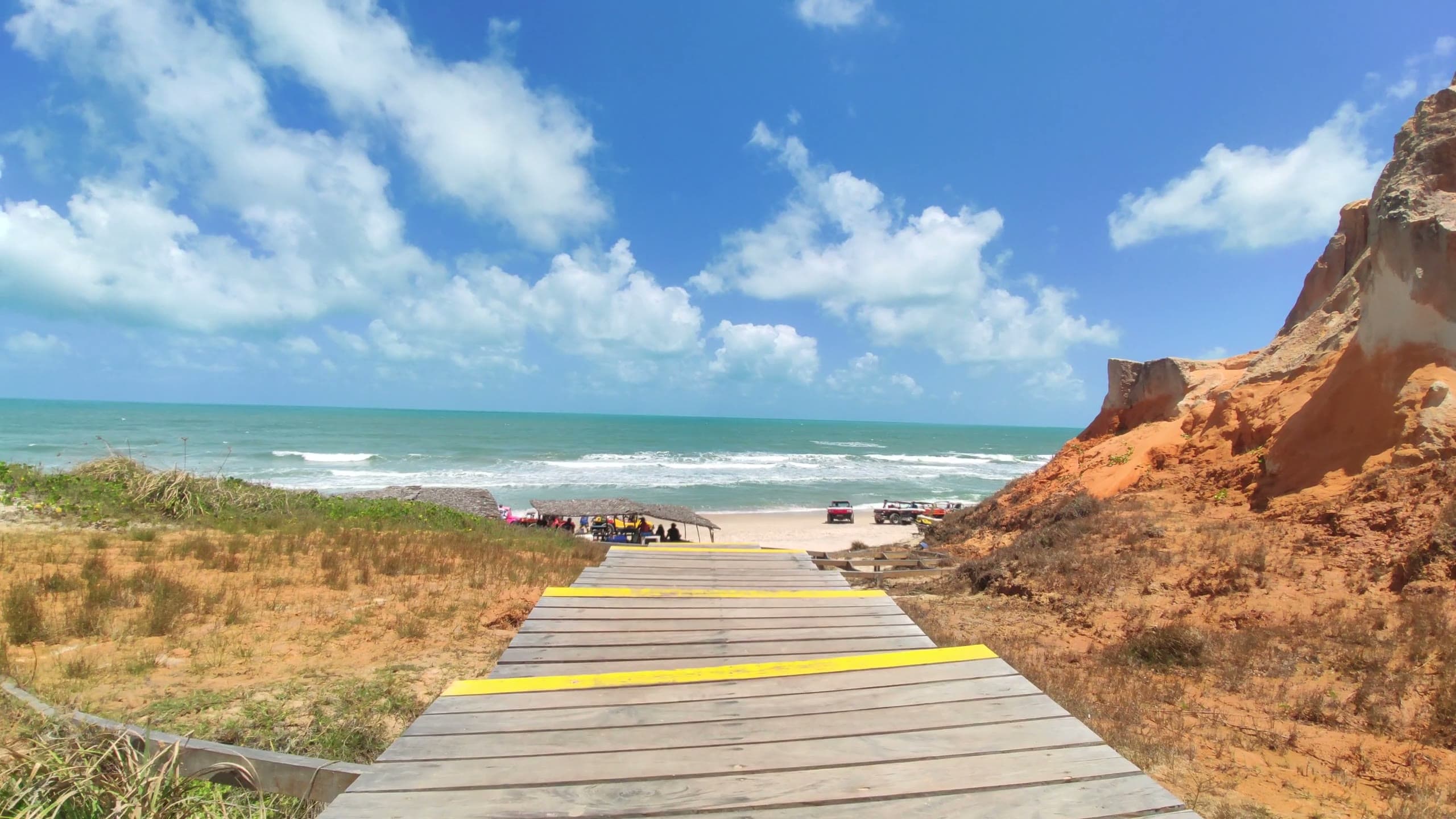 First-person view walking down a wooden boardwalk with yellow-striped steps toward a sunny beach, dune buggies and the open sea.