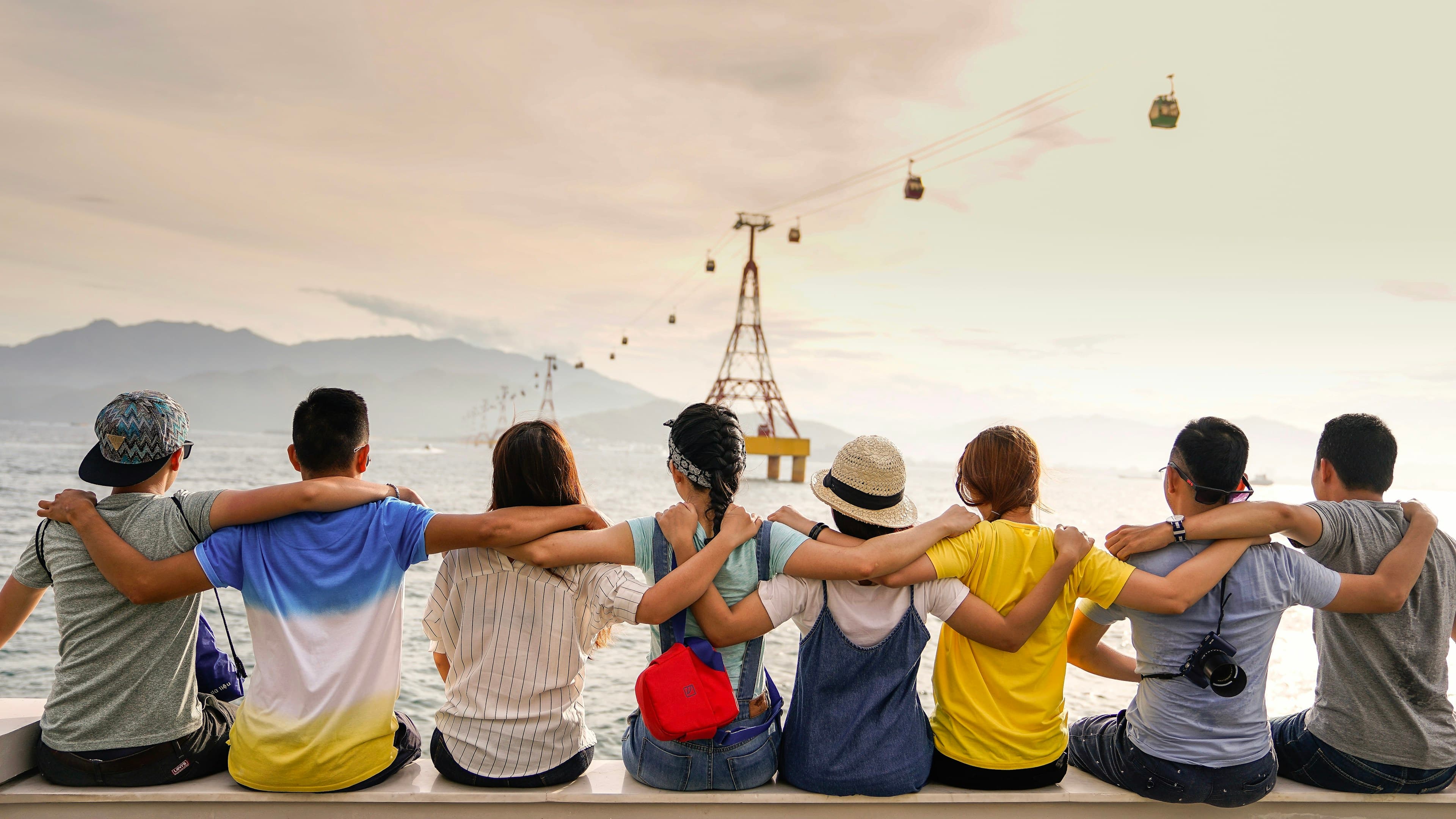 A group of friends sit with arms around each other, looking out at mountains and cable cars.