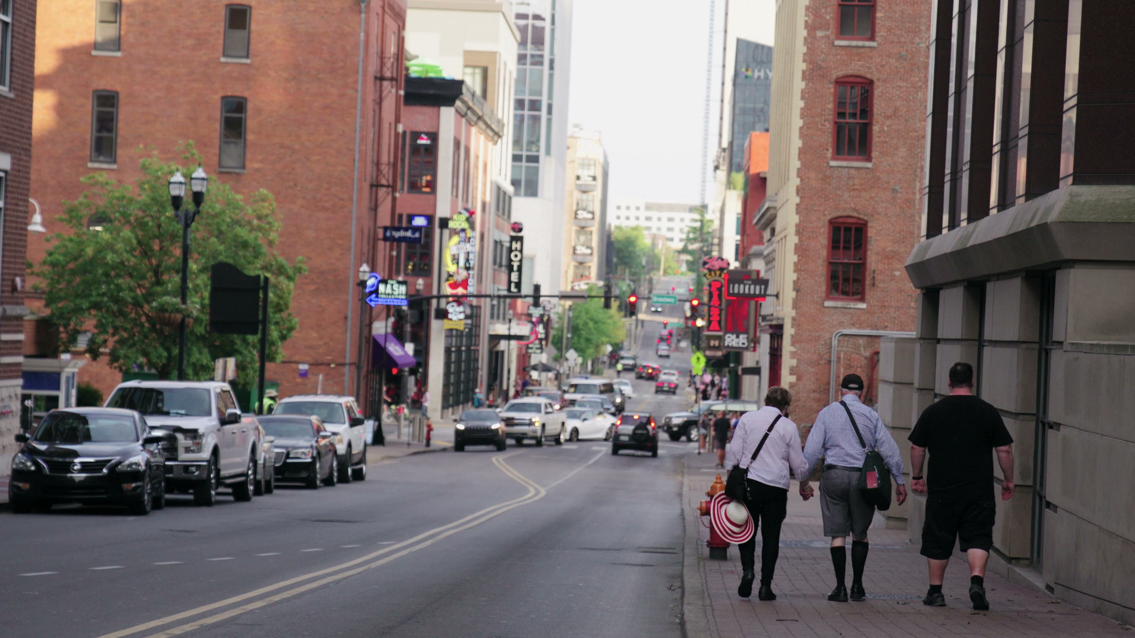 Three people walk away from the camera down a sunlit downtown sidewalk lined with brick buildings.