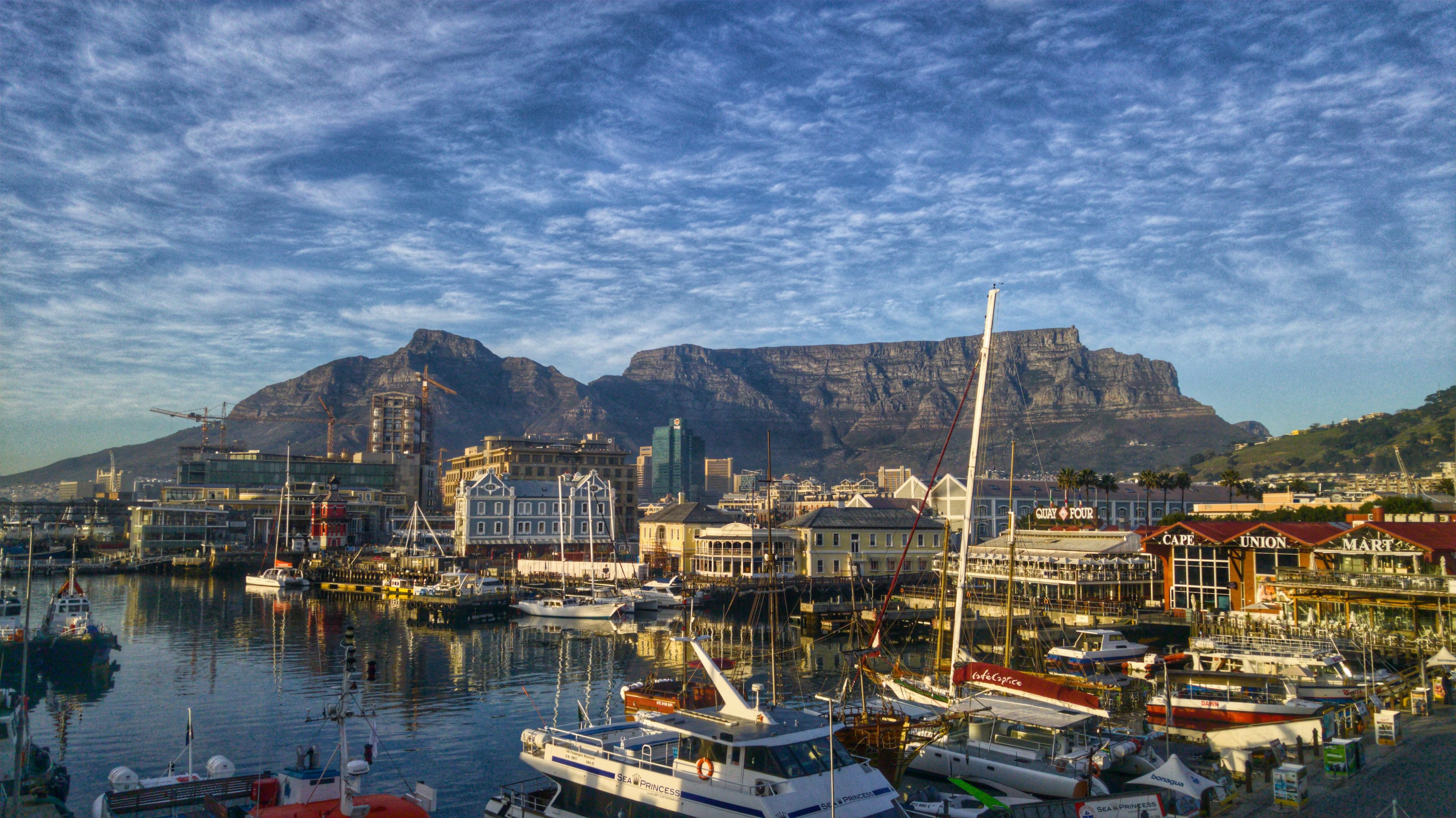 Cape Town's harbor: sailboats and yachts moored below waterfront buildings, with Table Mountain behind.
